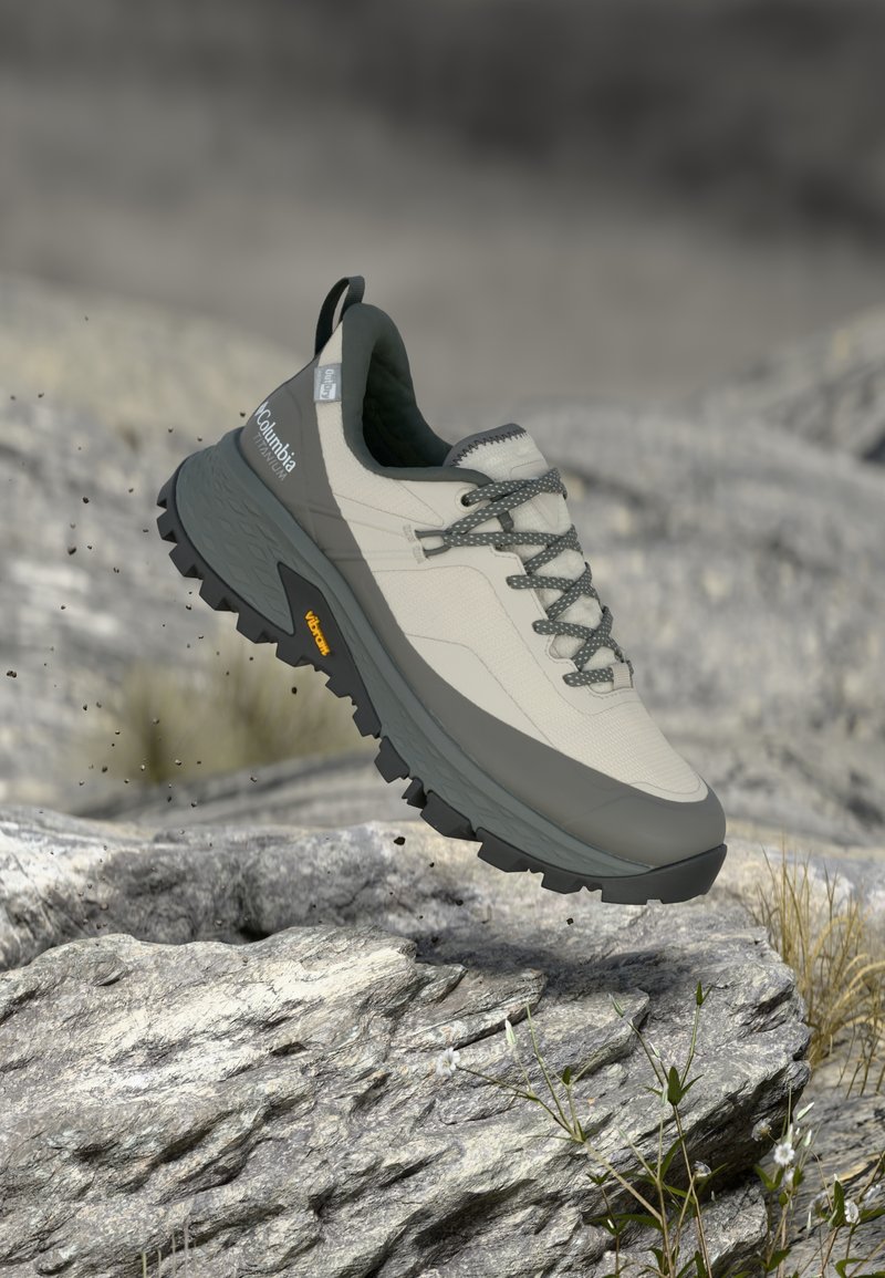 Chaussure d'extérieur beige clair avec des accents gris, surface texturée et semelle noire robuste. Dotée de lacets et d'une languette à l'arrière du talon.