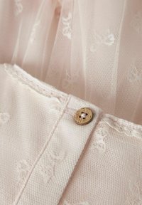 Vêtement rose clair avec une superposition de dentelle texturée. Présente un bouton en bois avec un design de fleur et des coutures délicates le long du bord.
