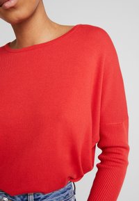 Roter Strickpullover mit weitem Ausschnitt und gerippten Manschetten, mit einem entspannten Schnitt und glatter Textur, kombiniert mit hoch taillierter Jeans.