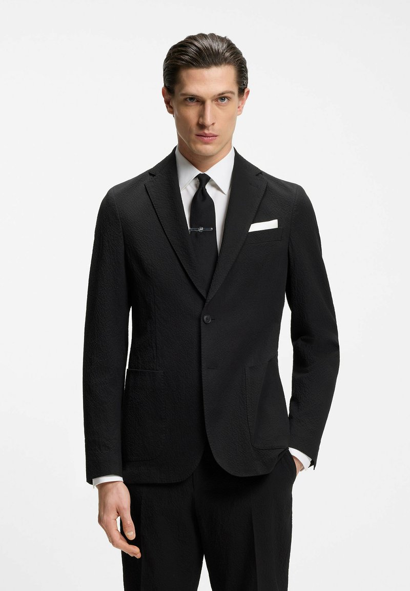 Homme en costume noir texturé, chemise blanche, cravate noire avec pince et pochette blanche, debout devant un fond blanc uni.