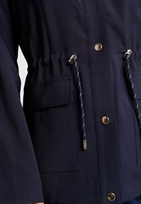 Gros plan d'une veste bleu marine avec poches avant, boutons-pression métalliques et cordons réglables à la taille.