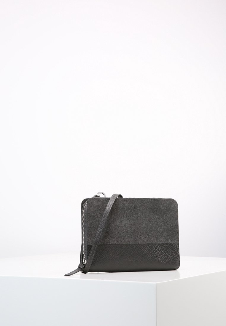 Petit sac bandoulière noir avec une partie supérieure en daim et une partie inférieure en cuir, posé sur un bloc blanc sur un fond clair uni.