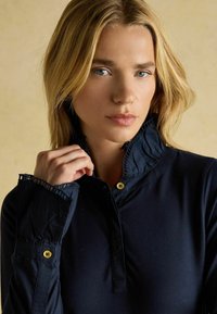 Tmavomodrá košeľa s dlhými rukávmi, volánovým golierom a manžetami, s doplnkami zo zlatých gombíkov. Hladká látka s priliehavým strihom.