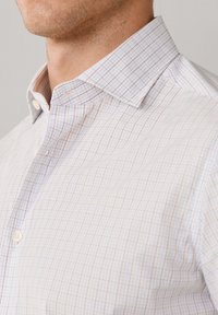 Camicia bianca con motivo a griglia blu e rossa. Presenta un colletto classico e una chiusura frontale con bottoni. Tessuto di cotone testurizzato.