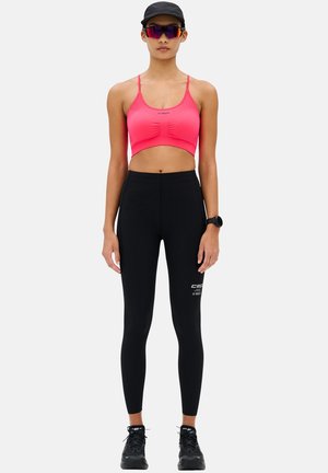 Athletic person wearing a pink sports bra, black leggings, black cap, sunglasses, watch, and black running shoes standing against white background.