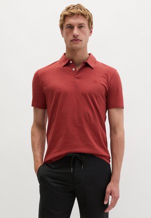 Mann trägt ein rostrot-kurzes Polohemd und schwarze Hose mit Kordelzug und steht vor einem einfarbigen hellen Hintergrund.