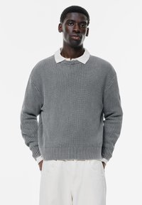 Maglione grigio a maglia con texture a coste, colletto polo bianco a contrasto, fit ampio e maniche lunghe. Indossato con pantaloni chiari.