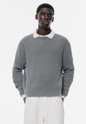 Pulover gri tricotat cu textură în dungi, guler de polo alb în contrast, croială lejeră și mâneci lungi. Purtați cu pantaloni de culoare deschisă.