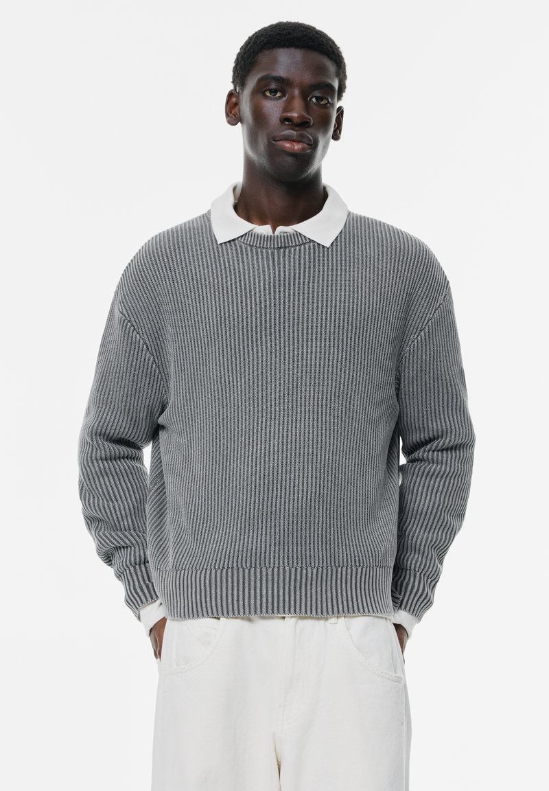 Maglione grigio a maglia con texture a coste, colletto polo bianco a contrasto, fit ampio e maniche lunghe. Indossato con pantaloni chiari.