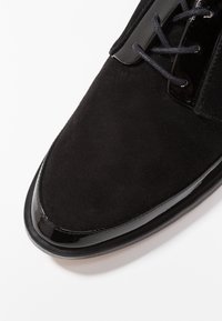 Czarne zamszowe buty z błyszczącą, lakierowaną czubkiem. Posiadają czarne sznurówki i gładkie wykończenie, podkreślające elegancki design.