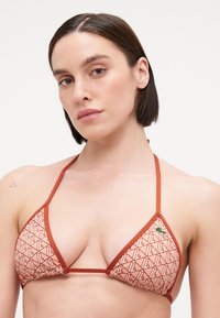 Triangular bikini top in coral with geometric pattern and contrasting orange trim. Features thin straps and a small green logo detail.