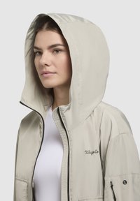 Beige hooded jacket with a zip front, featuring a smooth texture, side pockets, and subtle logo detail on the chest and sleeve.