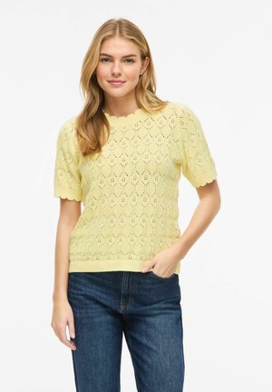 Jonge vrouw die een geel gebreid topje met korte mouwen en gegolfde randen draagt, gecombineerd met een blauwe spijkerbroek, staand met één hand in haar zak.