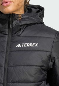 Primo piano di una persona che indossa un giubbotto imbottito nero con cappuccio, caratterizzato da un evidente logo bianco "TERREX" sul petto.