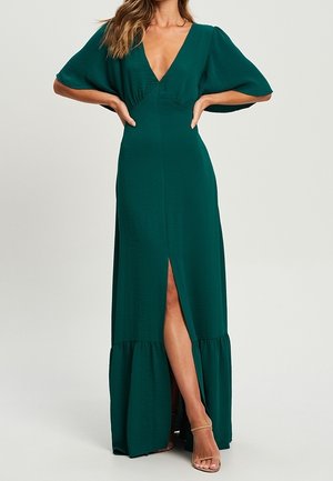 Woman wearing a dark green maxi dress with short puff sleeves, deep V-neck, front slit, and ruffled hem, paired with nude strappy heels.