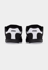 Ζευγάρι μαύρων και λευκών παπουτσιών Etnies, εμφανιζόμενα από πίσω, με έμφαση στο λογότυπο της μάρκας στην περιοχή της φτέρνας, πάνω σε απλό φόντο.