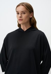 Felpa nera con cappuccio realizzata in tessuto morbido, con una vestibilità ampia e maniche lunghe. Design minimal con una texture liscia e opaca.