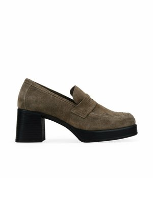 Mocasines de ante marrón con tacón cuadrado, punta redondeada y detalles de costura, que presentan una correa en el empeine y una suela negra texturizada.
