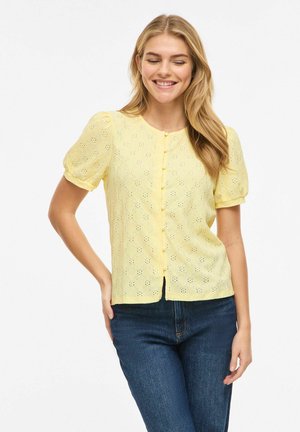 Femme souriante aux yeux fermés, portant un chemisier jaune à œillets boutonné et un jean bleu foncé, debout avec une main sur la hanche.