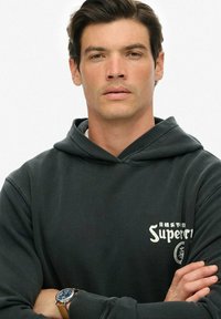 Joven con cabelo oscuro que lleva una sudadera negra de Superdry y un reloj en la muñeca, con los brazos cruzados, mirando hacia adelante frente a un fondo sencillo.