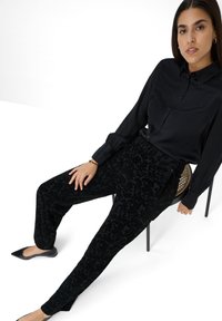 Schwarze Bluse mit Knopfverschluss und elastischen Bündchen, kombiniert mit einer strukturierten schwarzen gemusterten Hose. Modell sitzt auf einem Stuhl. Schwarze spitze Ballerinas.