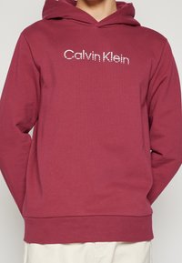 Burgundowa bluza z kapturem z kieszenią kangurką z przodu, z regulowanym kapturem i białym logo Calvin Klein na klatce piersiowej. Materiał mieszanki bawełny.