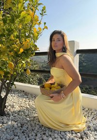 Femme en robe jaune agenouillée près d'un citronnier, tenant un panier de citrons sur une terrasse avec vue sur la montagne en arrière-plan.