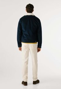 Giacca di jeans blu navy con colletto in finta pelliccia bianca, abbinata a pantaloni beige e scarpe marroni, vista da dietro.