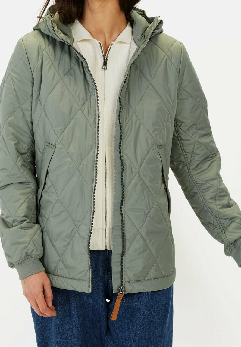 Personne portant une veste matelassée vert clair avec une fermeture éclair à l'avant, associée à un pull blanc zippé et un jean bleu.