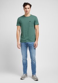 Grønn heathered kortermet skjorte med brystlomme, kombinert med lyseblå jeans og grå sneakers, stående mot en lys bakgrunn.