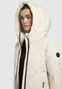 Giacca trapuntata in crema con imbottitura, colletto alto, cappuccio oversize e zip nera. Presenta una patch con logo sul maniche.