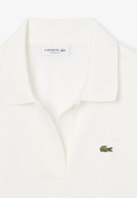 Vit Lacoste pikétröja med kort krage, prydd med en grön broderad krokodil-logga på vänster bröst. Tillverkad av strukturerad bomullstyg.