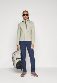 Giacca softshell grigia chiara con tasca sul petto con zip, pantaloni blu e uno zaino grigio chiaro e nero. Accompagnati da occhiali da sole e scarpe da trekking.