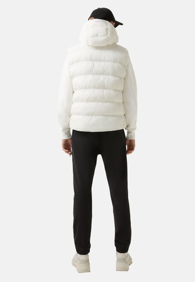 Witte gewatteerde vest met hoge kraag, gedeeltelijk gesloten capuchon, gecombineerd met zwarte joggingbroek en lichtgekleurde sneakers.