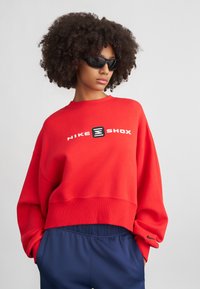 Červená skrátená mikina s bielym logom "NIKE SHOX", s rebrovanými manžetami a voľným strihom. Kombinovaná s námořníckymi nohavicami s malým akcentom Nike.