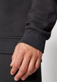 Černý svetr s manžetou s žebrovanou texturou, viděný s rukou položenou na oděvu. Tkanina se zdá být měkká a odolná.