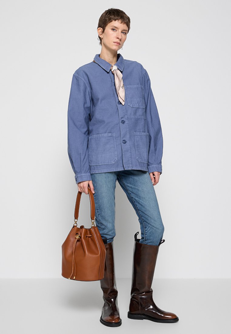 Blaue Baumwolljacke mit einem Knopfverschluss, zwei Fronttaschen, kombiniert mit blauen Jeans, braunen Leder-Stiefeln bis zum Knie und einer braunen Bucket Bag.