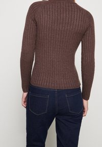 Bruine geribbelde trui met lange mouwen en een getailleerde pasvorm, gecombineerd met donkerblauwe jeans met twee achterzakken.