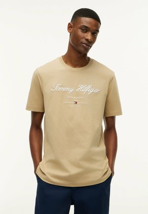 Ung mand iført en beige Tommy Hilfiger t-shirt med logo og tekst, stående med hænderne i lommerne mod en ensfarvet hvid baggrund.
