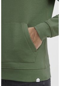 Green cotton sweatshirt with a kangaroo pocket and ribbed cuffs. Features a small hexagonal brand tag at the bottom edge.