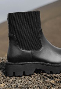 Botín de cuero negro hasta el tobillo con un upper de elastano acanalado, suela gruesa y texturizada, y detalles de costura en contraste.