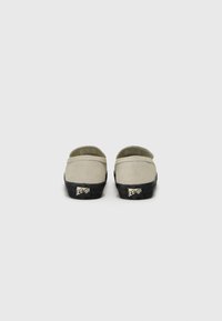 Beige slip-on shoes with a textured black sole. The back features a logo and a cushioned heel for comfort. Simple design, minimal detailing.