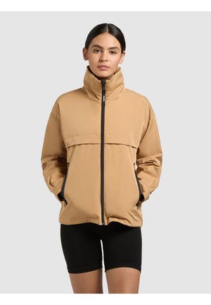 Nő, aki bézs színű, magas nyakú cipzáras dzsekit és fekete rövidnadrágot visel, zsebébe tett kézzel áll, egyszínű háttér előtt.