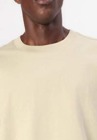 T-shirt beige clair à col rond, fabriqué en coton doux, avec une texture lisse et sans motifs ni accents visibles.