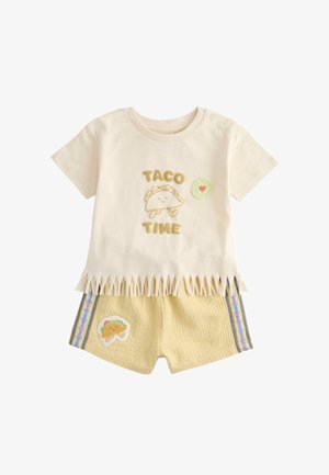 Kremowy strój dla malucha z napisem „Taco Time”, uśmiechniętą grafiką taco na koszulce, frędzlowanym dołem oraz pasujące szorty z naszywką taco i paskiem w paski po bokach.