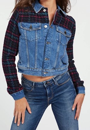 Femme portant une veste en jean courte avec des manches en maille à carreaux associée à un jean bleu taille haute.