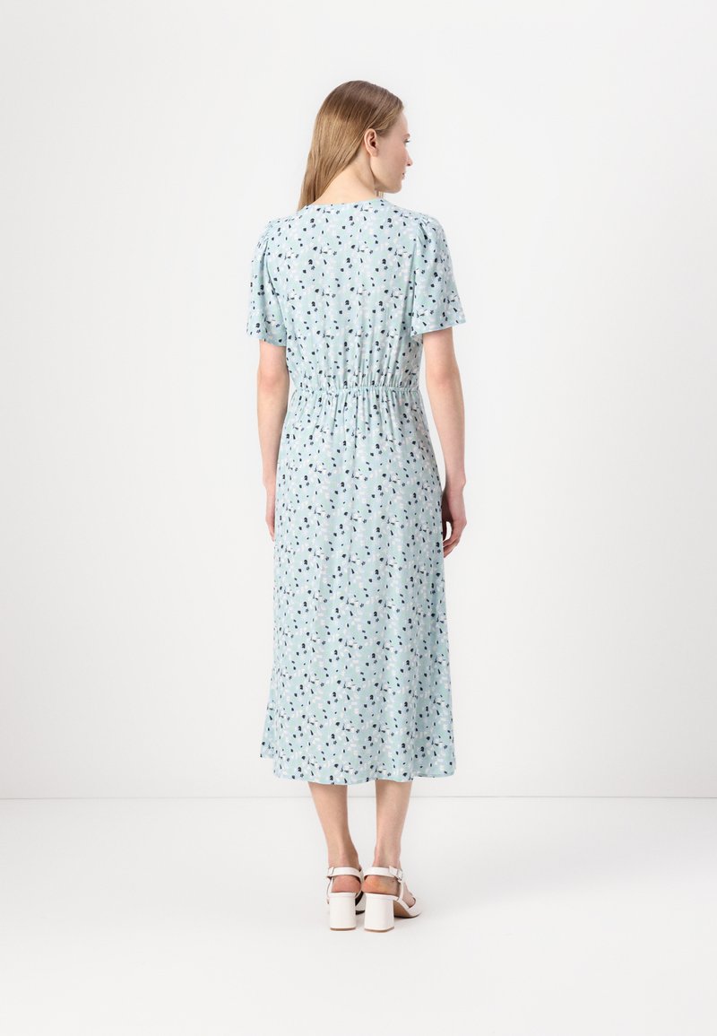 Sukienka maxi w jasnoniebieskim materiale z kwiatowym wzorem, elastycznym pasem, krótkimi rękawami i skromnym dekoltem, zestawiona z białymi sandałami na obcasie.