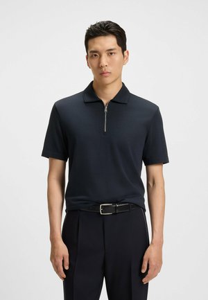 Polo shirt blu navy a maniche corte con colletto zip, realizzato in tessuto liscio. Indossato con pantaloni neri su misura e una cintura nera.