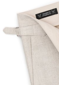 Tissu beige clair avec un motif chevrons et une étiquette cousue portant l'inscription « TAILORED TO FIT PERFECTLY », fixée par un fermoir métallique.