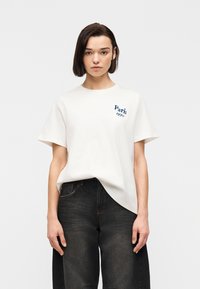 Νεαρή γυναίκα που φοράει λευκό μπλουζάκι με την επιγραφή «Παρίσι 1970» και άνετο μαύρο τζιν, στέκεται μπροστά σε απλό φωτεινό φόντο.